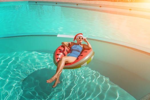 A happy woman in a blue bikini, a red and white Santa hat and sunglasses poses in the pool in an inflatable circle with a watermelon pattern, holding a glass of wine in her hands. Christmas holidays concept