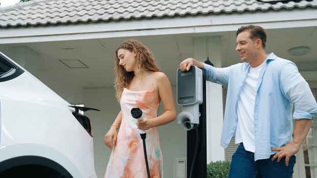 Happy and lovely couple with eco-friendly conscious recharging electric vehicle from EV charging station. EV car technology utilized as alternative transportation for future sustainability. Synchronos