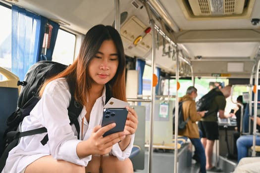 Happy Asian female tourist holding credit card and using smartphone while commuting by bus.