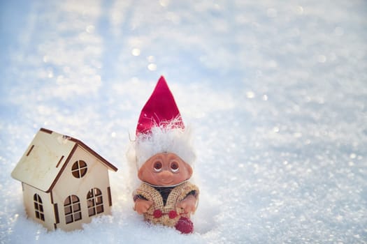 Cute little funny dwarf and small wooden house in snow in winter landscape. Wizard on snowy background. Christmas and New Year
