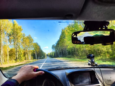view from a car windshield of natural landscape with road, green trees and blue sky in summer or spring time. Hand of woman on steering wheel. Female traveler driving alone on trip or journey
