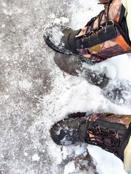 Feet of Hunter or fisherman in big warm boots on winter day on snow. Top view. Fisherman on the ice of a river, lake, reservoir on spring day with melting ice. Dangerous fishing in autumn