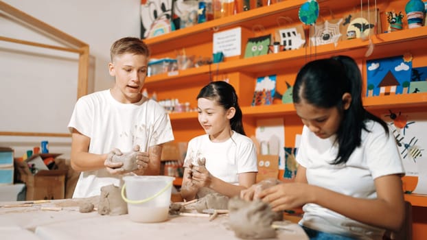 Happy diverse highschool student modeling clay at pottery workshop. Cute children with mixed raced working and making cup of clay by using carving equipment at art lesson. Creative class. Edification.