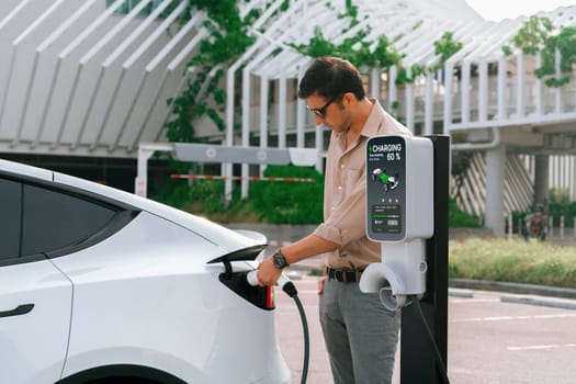 Young man put EV charger to recharge electric car's battery from charging station in city commercial parking lot. Rechargeable EV car for sustainable environmental friendly urban travel. Expedient