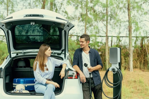Lovely young couple recharging battery for electric car while relaxing and sitting on the trunk during road trip travel EV car in autumnal forest. Eco friendly travel on vacation during autumn. Exalt