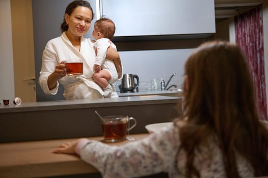 Authentic portrait of a young woman carrying her kid and having coffee in the morning at home with her daughter. Happy young family enjoying breakfast together.