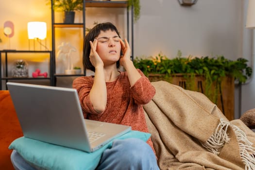 Tired young woman freelancer sitting on sofa using laptop, suffering from headache problem tension and migraine, stress. Caucasian girl feeling bored stretching arms sitting on orange sofa at home.