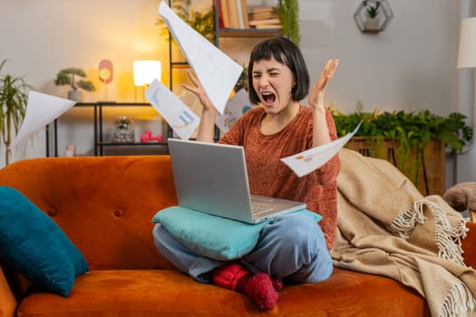 Angry young Caucasian woman freelancer with paperwork using laptop computer on sofa. Upset girl in casual clothes throwing documents in air closing netbook and looking away in living room at home.
