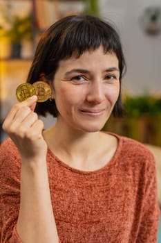 Portrait of smiling woman holding two golden bitcoins BTC, looking at camera. Happy surprised girl stock trader showing thumbs up sitting on sofa in living room at home. Increasing wealth. Vertical