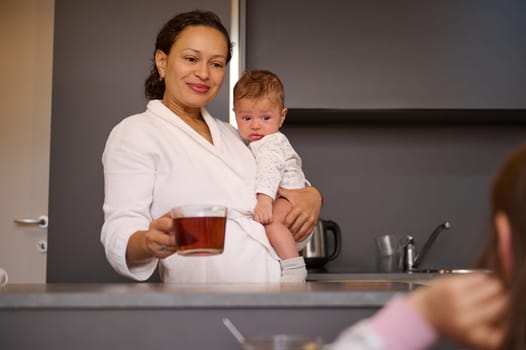 Smiling multi ethnic pretty woman, young mother carrying her lovely newborn baby boy, holding a cup of hot tea while standing at kitchen counter, enjoying a happy day with her kids at home