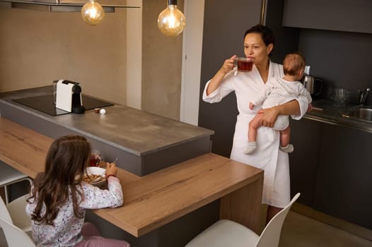 Young woman drinking tea, holding her baby, standing in the home kitchen interior. On the foreground, cute schoolgirl, adorable daughter eating healthy cereals with milk for breakfast in the morning