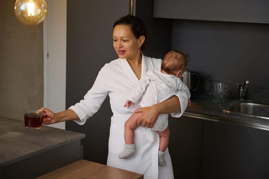 Beautiful multi ethnic young woman in white bathrobe, a loving happy carrying her baby and drinking tea in the morning for while breakfast time. People. Food and drink. Maternity lifestyle