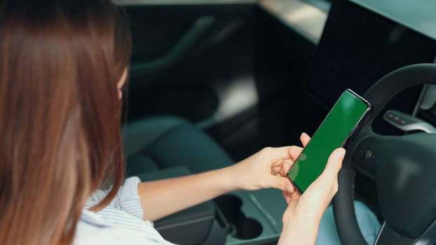 Holiday vacation road trip with environmental-friendly car concept. Eco-conscious woman on driver seat holding blank copyspace green screen smartphone for EV battery status. Exalt