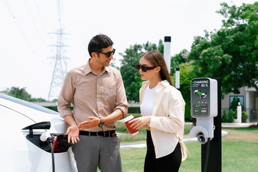 Young couple with coffee pay for electricity while recharge EV car battery at charging station connected to electrical power grid tower as electric industrial for eco friendly car travel. Expedient