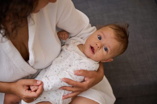 View from above portrait of a Caucasian adorable blue eyed baby boy 5 months old, dressed in white baby bodysuit, smiling looking at camera, nestling in his mother's arms. Babyhood and infancy concept