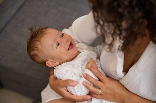 Adorable baby boy nestling in his mother's arms, smiles looking at his mom stroking, kissing, embracing and cuddling him. Young affectionate loving caring mother enjoys happy moments with her child