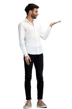 A young man on a white background in full growth shows his hand to the side.