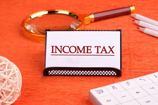 INCOME TAX text written on a white business card on a stand on an orange background, next to a magnifying glass, a calculator and pencils