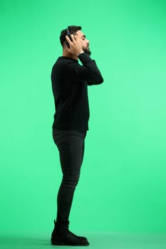 A man, in full height, on a green background, listens to music with headphones.