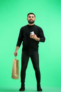A man, full-length, on a green background, with bags and a phone.