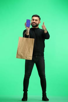 A man, full-length, on a green background, with bags, shows a phone.