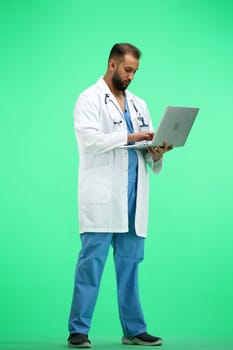 The doctor, in full height, on a green background, uses a laptop.