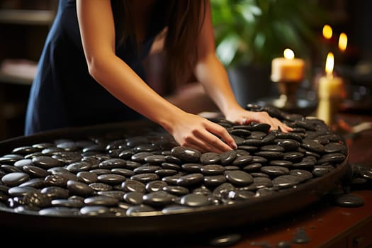 Woman stacks black massage stones in spa salon, black massage stones close-up in large wooden bowl.