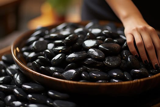 Woman stacks black massage stones in spa salon, black massage stones close-up in large wooden bowl.
