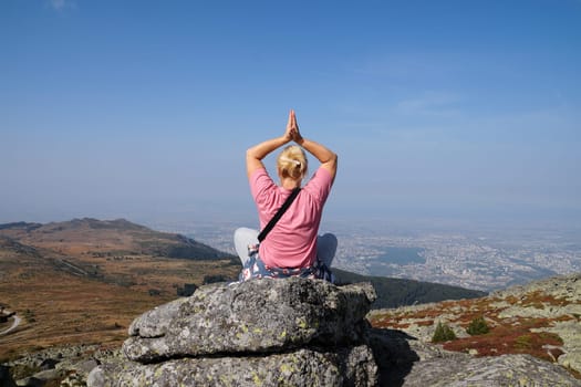 blonde woman meditates in a yoga pose with her arms raised above her head on the top of a mountain, back view