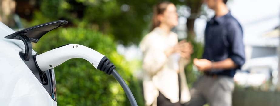 Young couple recharge electric car battery from charging station in green city park in springtime. Rechargeable EV car for sustainable environmental friendly urban travel lifestyle. Panorama Expedient