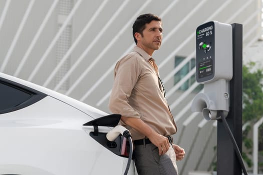 Young man put EV charger to recharge electric car's battery from charging station in city commercial parking lot. Rechargeable EV car for sustainable environmental friendly urban travel. Expedient