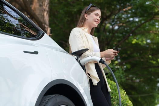 Young woman use smartphone to pay for electricity at public EV car charging station green city park. Modern environmental and sustainable urban lifestyle with EV vehicle. Expedient