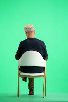 A man, full-length, on a green background, sitting on a chair.