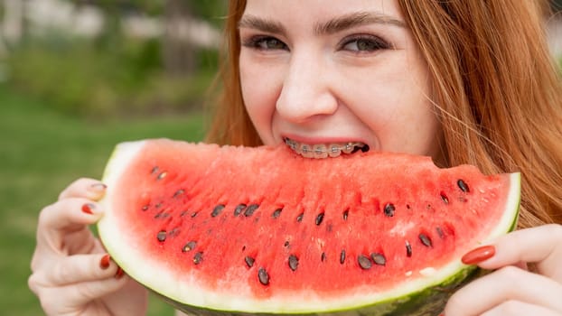 Close-up portrait of red-haired young woman with braces eating watermelon outdoors.