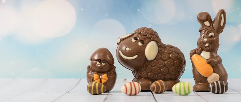Cute chocolate happy lamb, bunny, chicken in the shell and easter candy eggs around them stand on the right side on a white wooden table with copy space on the left, close-up side view.