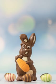 One chocolate happy bunny with an orange carrot in his hands and scattered easter candy eggs around him stands on a white wooden table with a little copy space on top, close-up side view.