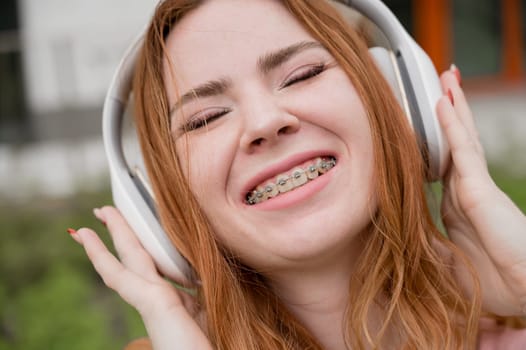 Portrait of a young red-haired woman with braces on her teeth listening to music on headphones outdoors