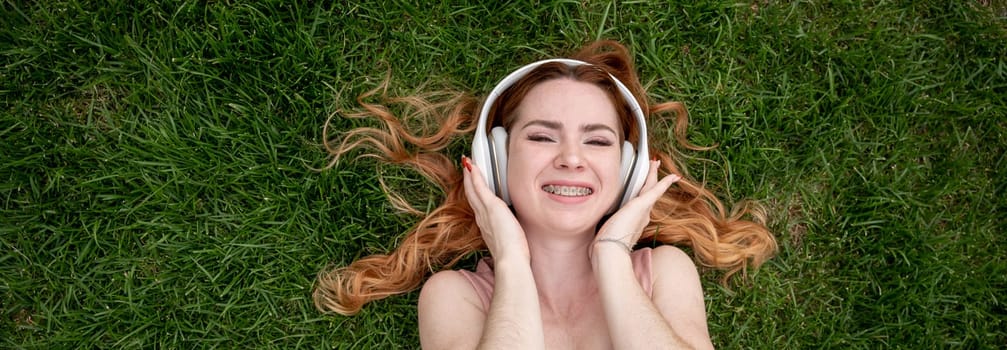 Top view of a young red-haired woman lying on the grass and listening to music on headphones. Widescreen