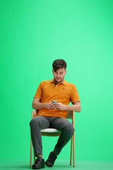A man, full-length, on a green background, sitting on a chair.