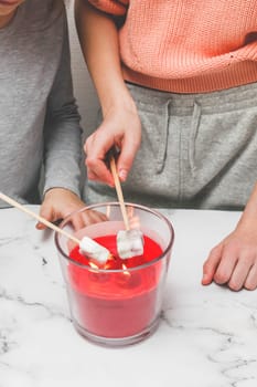 Two little girls in home clothes fry marshmallows over a candle fire, holding them on wooden skewers, standing at a light marble table, close-up side view.