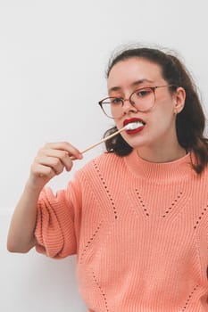 One beautiful Caucasian teenage girl in glasses and a peach sweater eats fried marshmallows on a skewer while standing against a white wall in the kitchen during the day, side view close-up.