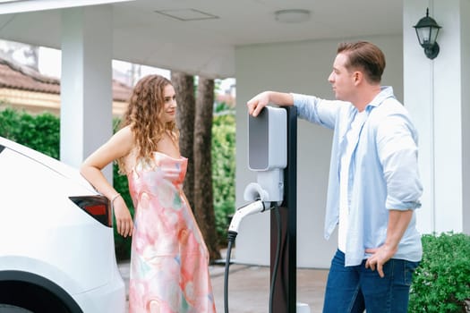 Happy and lovely couple with eco-friendly conscious recharging electric vehicle from EV home charging station. EV car technology utilized for residential home to future sustainability. Synchronos