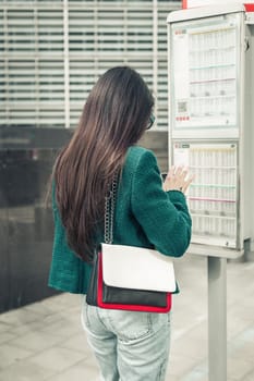 One young caucasian brunette girl with long flowing hair in a green jacket stands with her back at a bus stop near the schedule board, close-up side view.