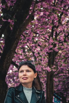 Portrait of one young beautiful caucasian girl in a leather jacket with a smile stands on a city street against the backdrop of flowering sakura trees, side view close-up.