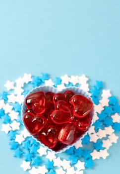 One cupcake pan with glass red hearts and candy sprinkles stars around it lies from below on a blue background with copy space on top, closeup flat lay. Valentine's day concept, gender party.
