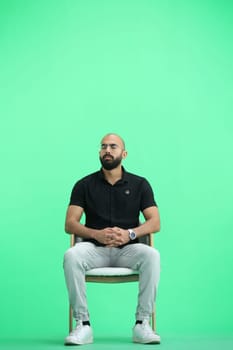 A man, on a green background, in full height, sitting on a chair.