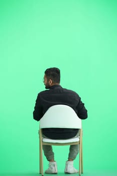 A man, on a green background, in full height, sitting on a chair.