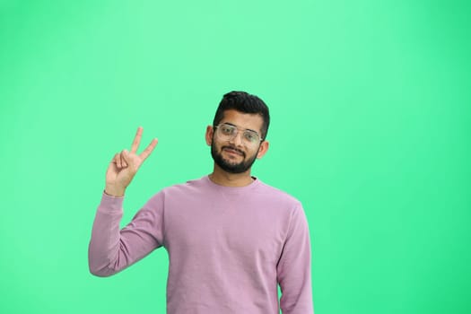 A man, on a green background, in close-up, shows a victory sign.