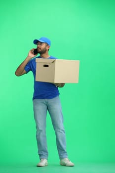 A male deliveryman, on a green background, in full height, with a box and a phone.
