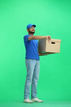 A male deliveryman, on a green background, full-length, with a box, shows his finger down.